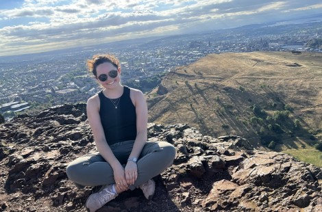 Andrea is sat cross-legged on Arthur's Seat in Edinburgh. It is a sunny day and she is smiling, wearing sunglasses.